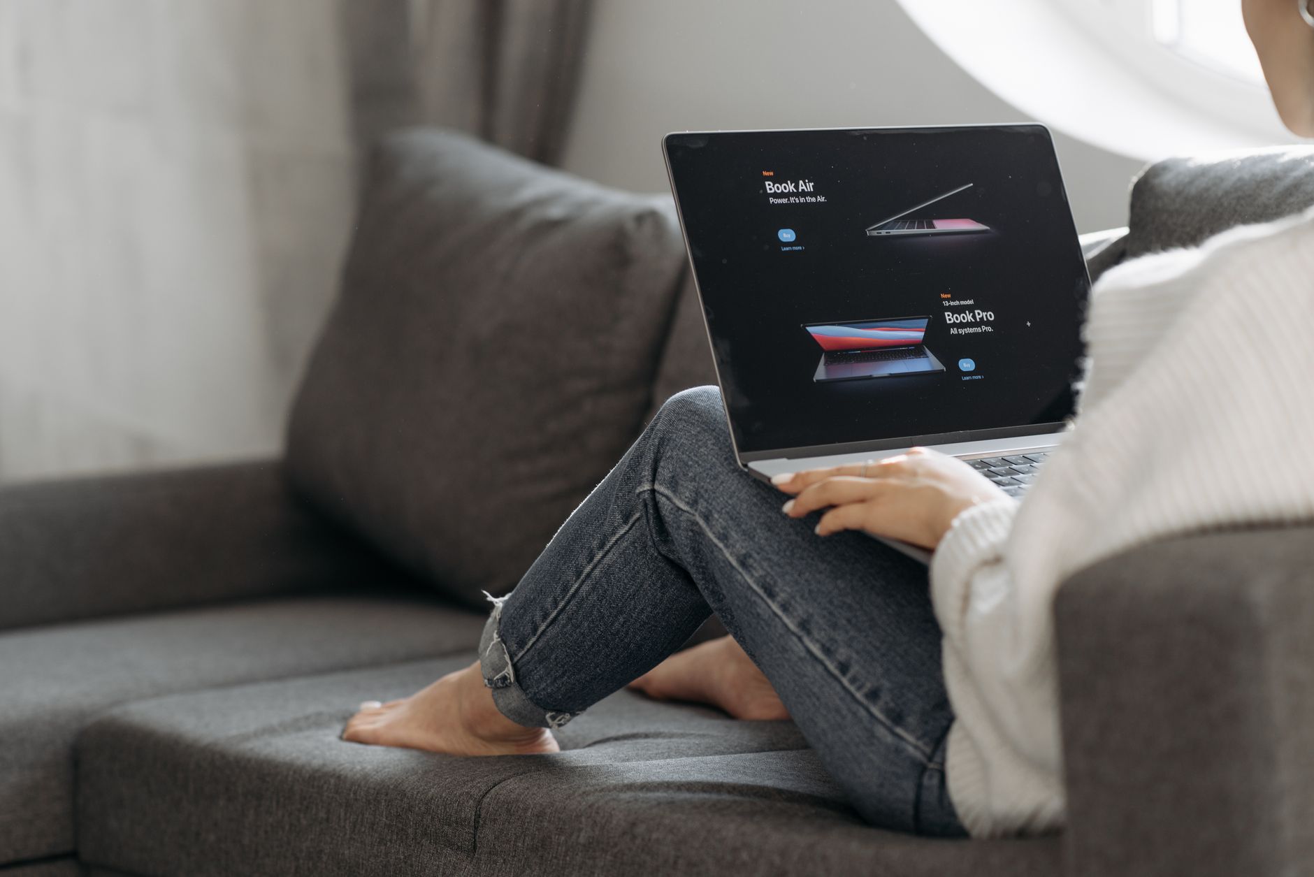 Une personne installée sur un canapé gris, un ordinateur portable sur les genoux, parcourt un site web depuis son salon
